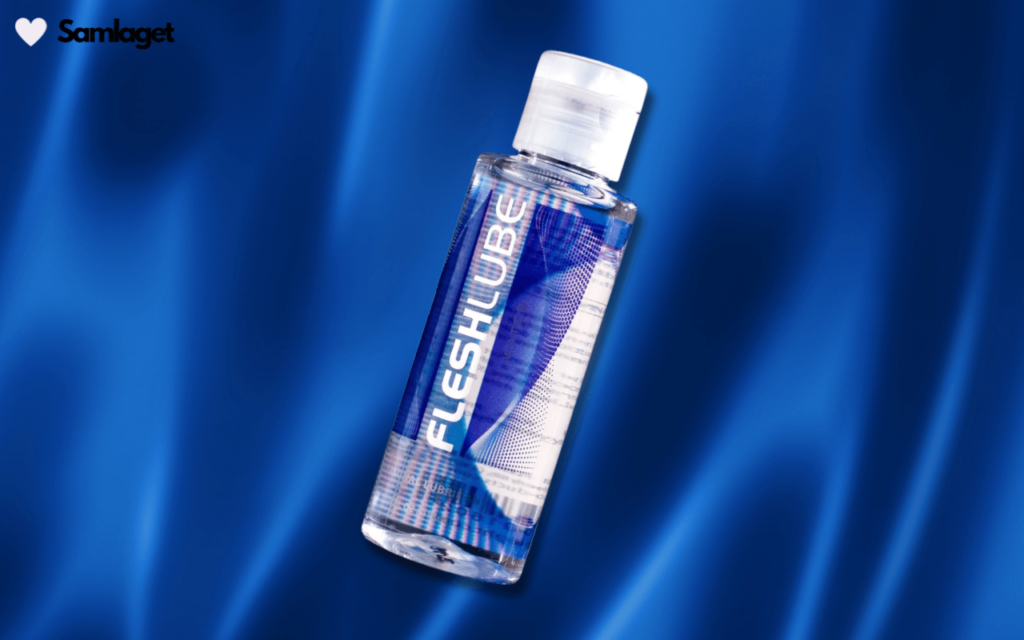 En genomskinlig flaska med glidmedel från varumärket Fleshlube, liggande snett på en blå silkesliknande tygbakgrund. Flaskan har en vit skruvkork och en etikett med blå och vita detaljer samt texten 'FLESHLUBE'.