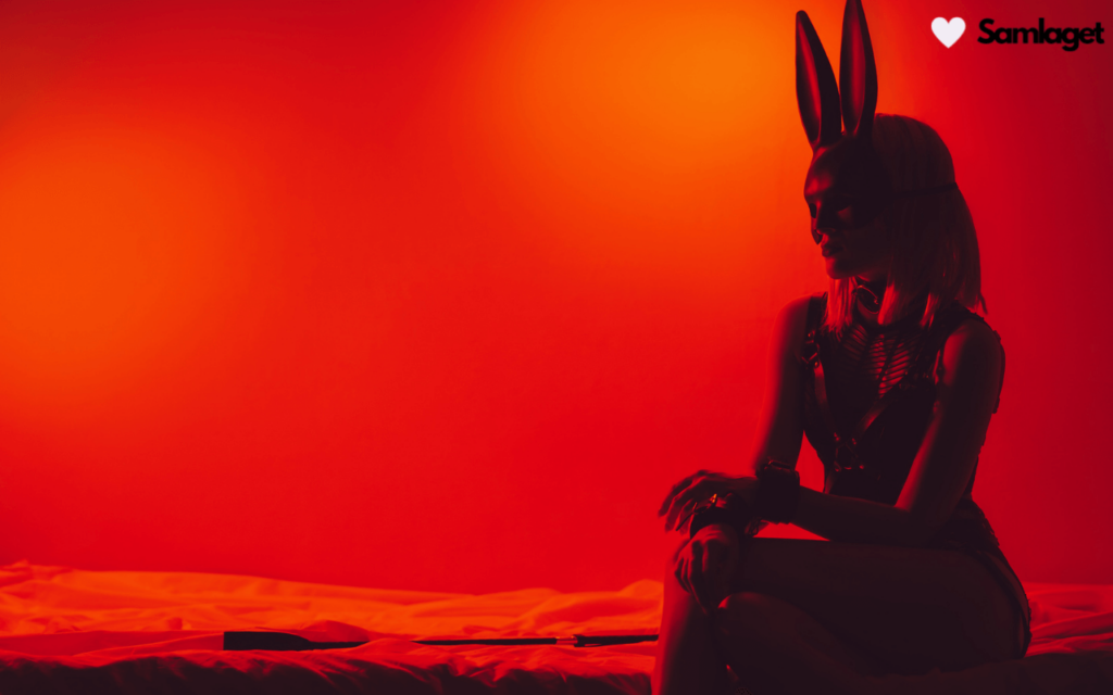 Kvinna i kaninmask och BDSM-utstyrsel sitter på säng med röd bakgrundsbelysning.